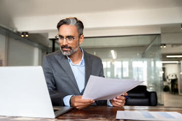 Focused Hispanic businessman with paperwork in office. Busy Latin or Indian male business man accountant analyst holding documents, work at laptop computer doing online trade market tech research. 