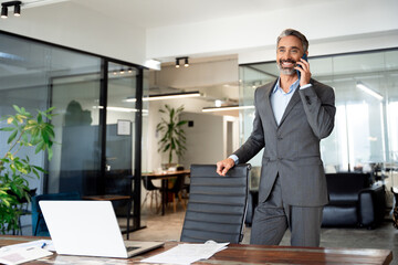 Middle aged Latin or Indian businessman having call on smartphone with business partners or clients. Smiling mature Hispanic man in relaxing pose talking by mobile cellphone at work in modern office. 