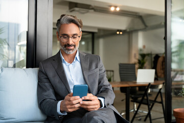 Close-up of smiling mature Latin or Indian businessman holding smartphone in office. Middle aged manager using cell phone mobile app. Digital technology application and solutions for business concept.