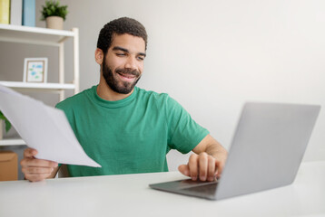 Smiling young man in green t-shirt happily reviewing documents while simultaneously working