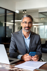 Vertical portrait of middle aged Hispanic business manager using cell phone mobile app. Smiling Latin or Indian mature man businessman holding smartphone sit in office working online on gadget.