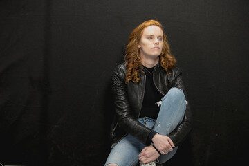 Young woman with red hair in black jacket and jeans poses in studio before a black background