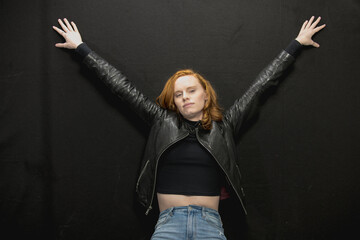 Young woman with red hair in black jacket and jeans poses in studio before a black background