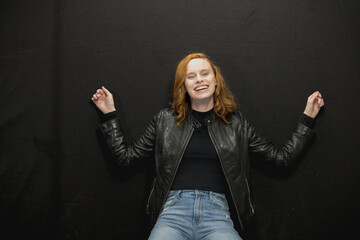 Young woman with red hair in black jacket and jeans poses in studio before a black background