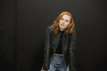 Young woman with red hair in black jacket and jeans poses in studio before a black background