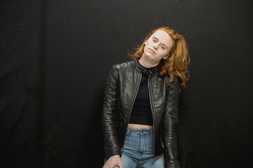 Young woman with red hair in black jacket and jeans poses in studio before a black background