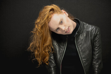 Young woman with red hair in black jacket and jeans poses in studio before a black background