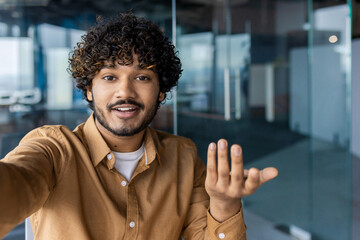 Charming young man with curly hair, dressed casually, extends his hand in greeting, inviting engagement in a bright office setting.