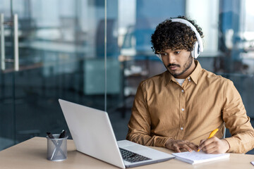 A young man in casual work attire and headphones focuses intently on his work at a laptop, taking...