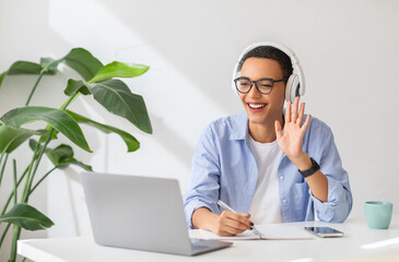 Glad young latin lady in headphones work, study at table, has video call, lesson, waving hand on...