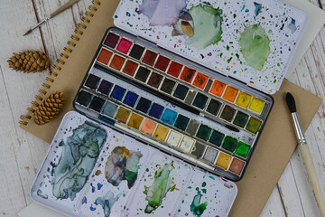 Large set of watercolor paints on a wooden table. View of the object from above. Accessories for the artist's drawing. Paints, sketchbook, brushes.