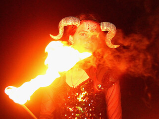 Selfie portraits of a red-haired woman in a shiny gothic outfit, with horns and burning torches (double staff fire prop). Mystical fire show at night.
