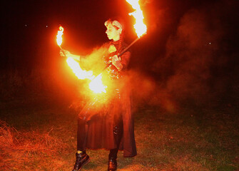 Selfie portraits of a red-haired woman in a shiny gothic outfit, with horns and burning torches (double staff fire prop). Mystical fire show at night.