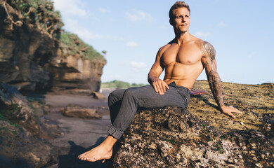Fototapeta na wymiar Muscular man in pants sitting on rock at seashore