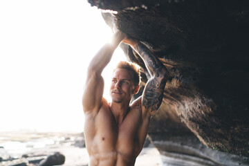 Muscular man with naked torso raising arms on rocky cliff on sunny day