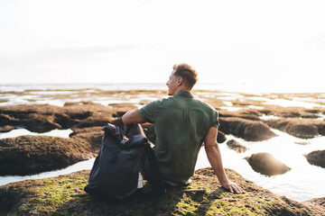 Unrecognizable male tourist sitting on rocky seashore in daytime with bag - Powered by Adobe