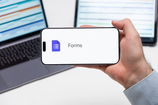 Forms application logo on the screen of smart phone in mans hand, laptop and tablet are on the table in the background, December 2023, Prague, Czech Republic.