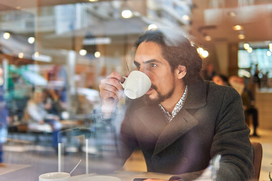 Through window view of thoughtful mature Latin businessman with long hair looking away while drinking coffee in a downtown cafe