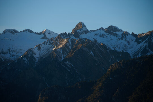 Early morning light bathes the rugged peaks of the Pyrenees in a warm glow, the snow-capped mountains contrasting against the clear blue sky.