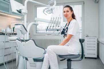 Portraait of sincerely smiling young dentist woman dressed white medical scrubs uniform sitting in modern dental clinic next to stomatology  chair. Health care and medicare industry concept image.