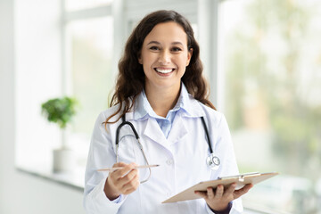 Cheerful young european woman doctor posing by window, holding clipboard