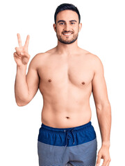 Fototapeta premium Young handsome man wearing swimwear smiling with happy face winking at the camera doing victory sign. number two.