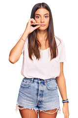 Young hispanic woman wearing casual white tshirt mouth and lips shut as zip with fingers. secret...