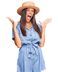 Young hispanic woman wearing summer hat celebrating mad and crazy for success with arms raised and...