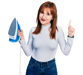 Redhead young woman holding electric steam iron surprised with an idea or question pointing finger with happy face, number one