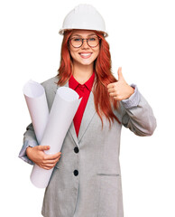 Young redhead architect woman wearing safety helmet holding blueprints smiling happy and positive, thumb up doing excellent and approval sign