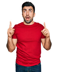 Fototapeta premium Hispanic man with beard wearing casual red t shirt amazed and surprised looking up and pointing with fingers and raised arms.
