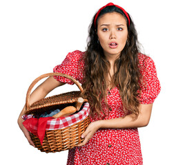 Young hispanic girl holding picnic wicker basket with bread clueless and confused expression. doubt...