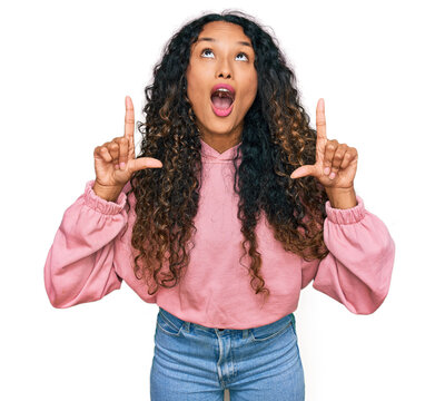 Young hispanic woman with curly hair wearing casual sweatshirt amazed and surprised looking up and pointing with fingers and raised arms.