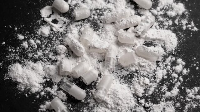 Heaps of white powder and scattered open medical capsules on a black table. Narcotic party and drug addiction concept. Rotation. Top view