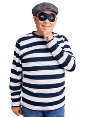 Senior handsome man wearing burglar mask and t-shirt looking confident at the camera smiling with crossed arms and hand raised on chin. thinking positive.