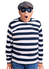 Senior handsome man wearing burglar mask and t-shirt afraid and shocked with surprise and amazed expression, fear and excited face.