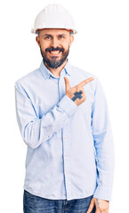 Young handsome man wearing architect hardhat cheerful with a smile of face pointing with hand and finger up to the side with happy and natural expression on face