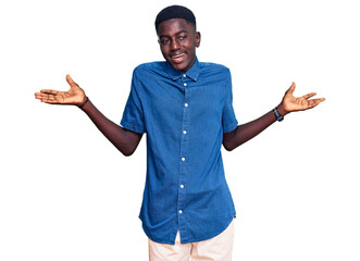 Naklejka premium Young african american man wearing casual clothes clueless and confused expression with arms and hands raised. doubt concept.