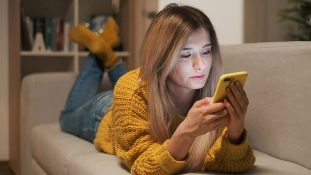 A pretty young woman using her smartphone on the couch, texting while relaxing. Attractive girl in yellow sweater using smart phone, laughing while lying on the sofa in evening.