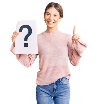 Beautiful caucasian woman with blonde hair holding question mark surprised with an idea or question pointing finger with happy face, number one