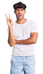 Young hispanic man wearing casual clothes smiling with happy face winking at the camera doing victory sign. number two.