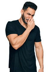 Hispanic man with beard wearing casual black t shirt smelling something stinky and disgusting, intolerable smell, holding breath with fingers on nose. bad smell