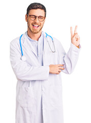 Handsome young man with bear wearing doctor uniform smiling with happy face winking at the camera doing victory sign. number two.