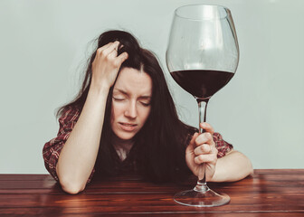 A young caucasian girl in a plaid shirt with tousled hair sits at a wooden table and holds a large glass of red wine.Concept of alcohol abuse, headache, alcoholism, hangover, depression and loneliness