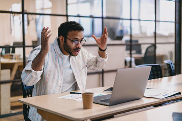 Anxious ethnic man with raised hands working with laptop in office