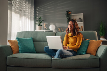 Woman using laptop and talking with a client on a phone while sitting on couch - Powered by Adobe
