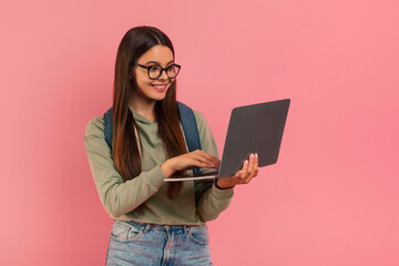 Smiling teenage girl wearing eyeglasses and backpack looking at laptop screen - obrazy, fototapety, plakaty