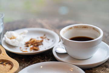 An after-meal scene with an empty coffee cup, a crumpled napkin, and croissant crumbs on a wooden tray and a white plate.