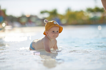 a little half a year old, a baby baby crawling on the water, smiling, outdoors, in a panama hat, in diapers, in the sun at the resort.