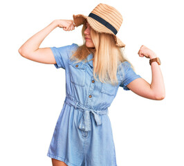 Young beautiful blonde woman wearing summer hat and dress showing arms muscles smiling proud. fitness concept.